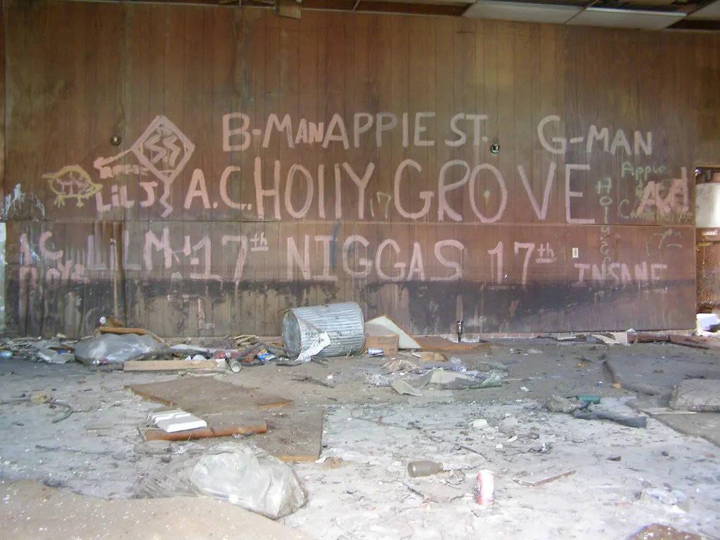 Abandoned room with graffiti-covered walls and debris across the floor beneath an incomplete suspended ceiling.