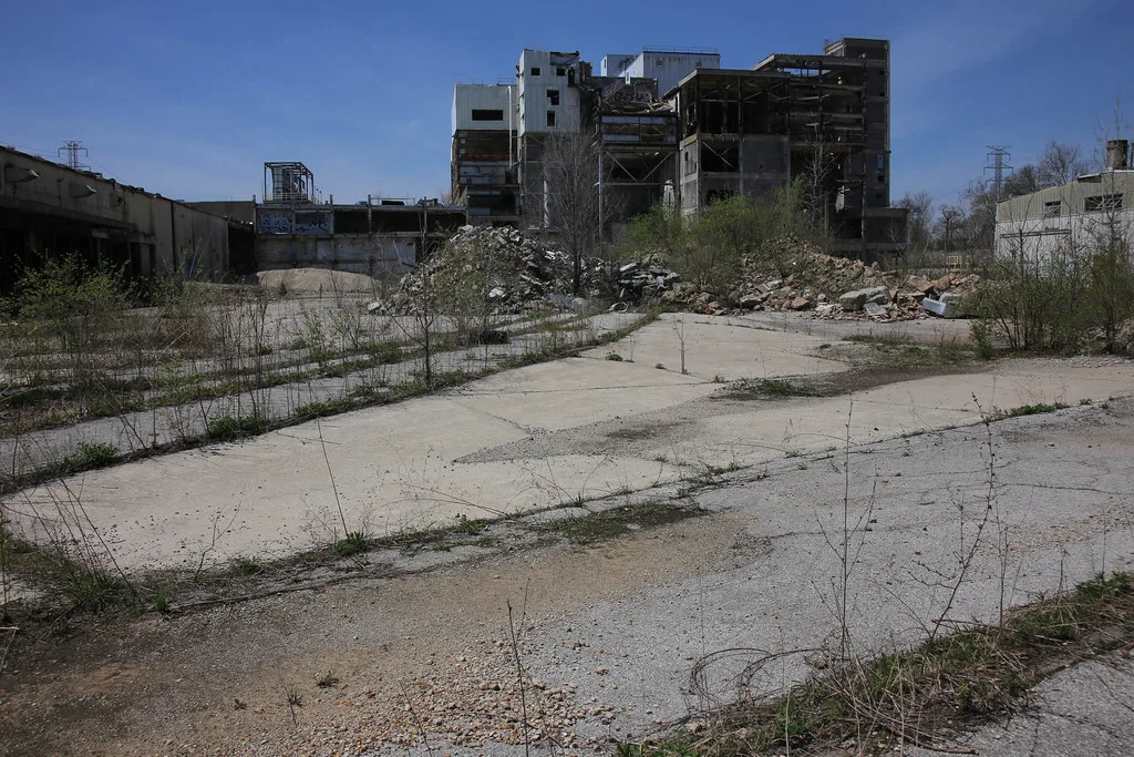 Partly dismantled abandoned factory behind a cracked concrete yard and rubble piles overgrown with plants.