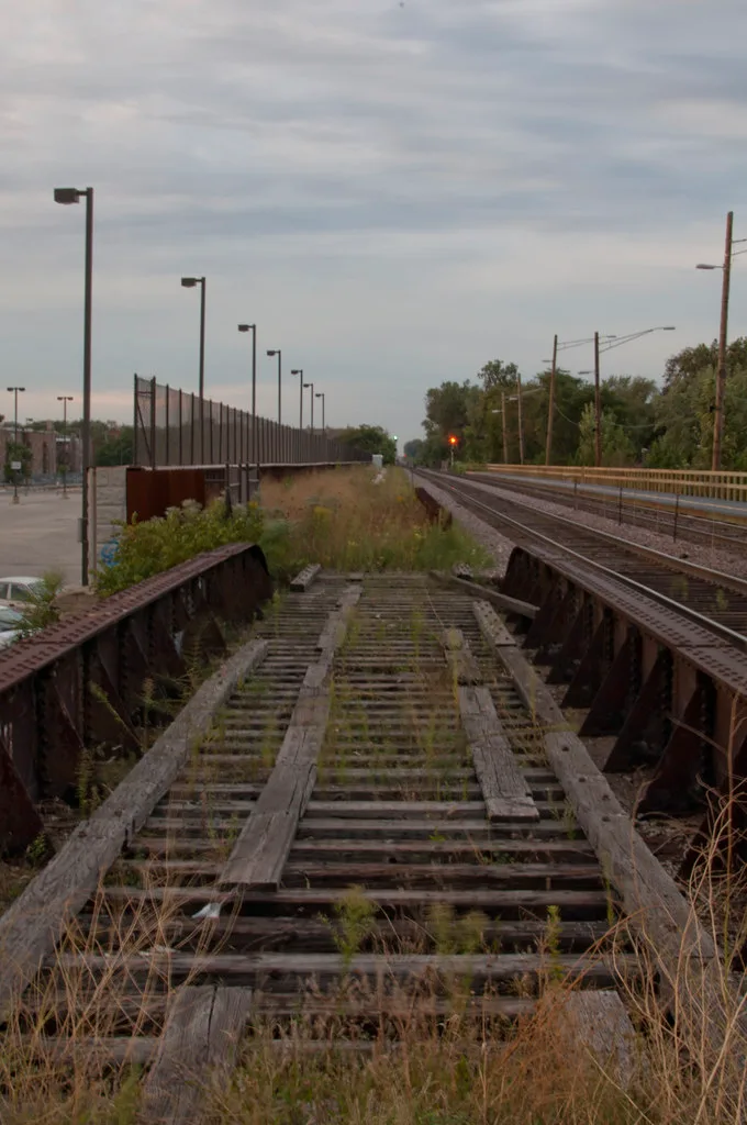 Disused railway bridge with bare ties and encroaching plants beside electrified tracks.