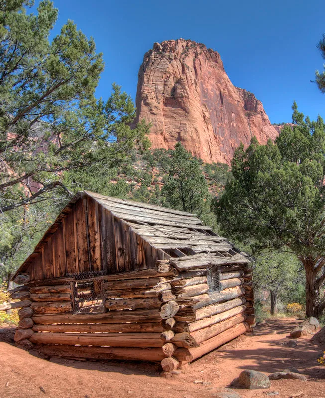 Log cabin with a stripped roof below a tall red cliff.
