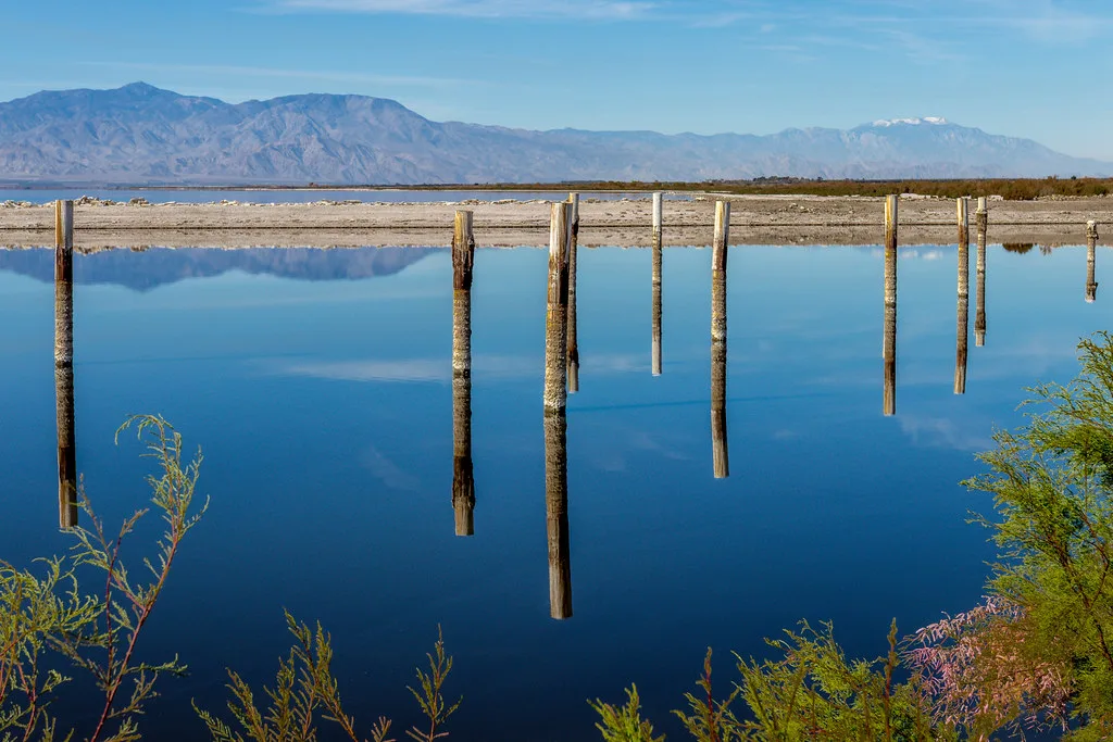Row of wooden piles without a dock reflected in still water before mountains.
