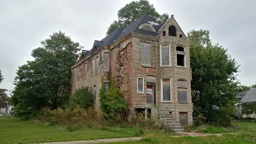 Abandoned house with a stone bay window, boarded or empty openings and missing render exposing brickwork.