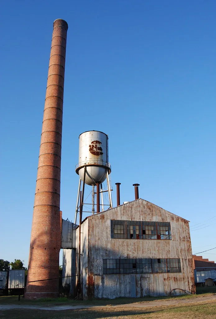 Disused industrial building with rusty metal siding beneath a tall brick chimney and water tower.