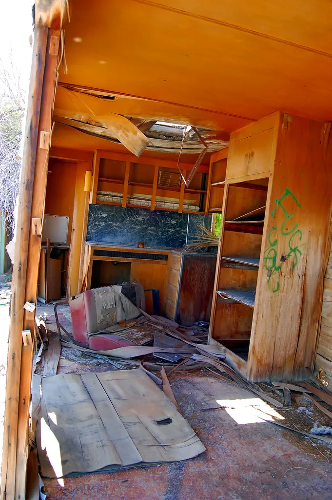 Abandoned interior with orange wooden cabinets, a broken ceiling and a torn seat, with boards and debris covering the floor.