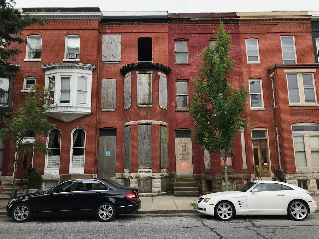 Neglected brick row houses with boarded openings and broken windows behind two parked cars.