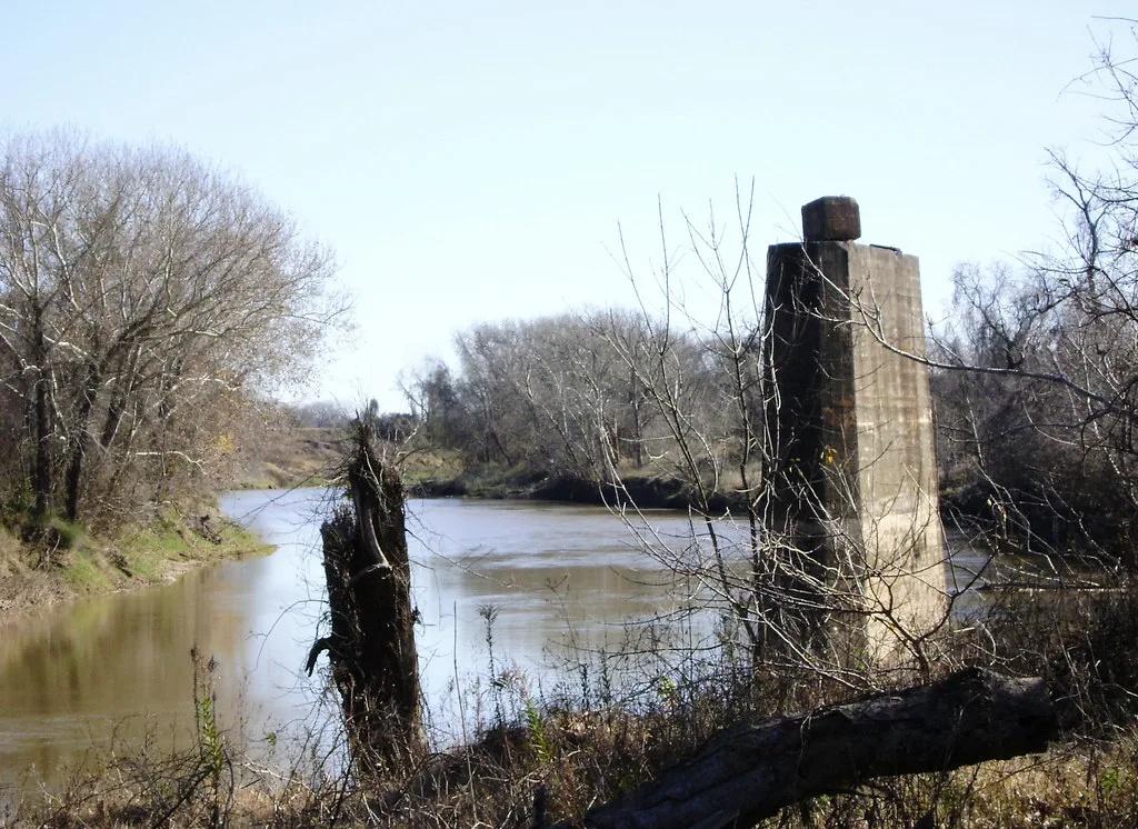 Old concrete bridge pier without a deck among trees beside a brown river.