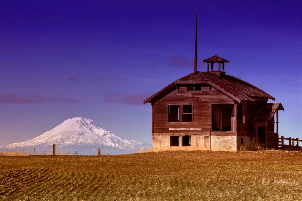 Abandoned old wooden building with a small bell tower and broken windows facing a field and a snow-covered mountain.