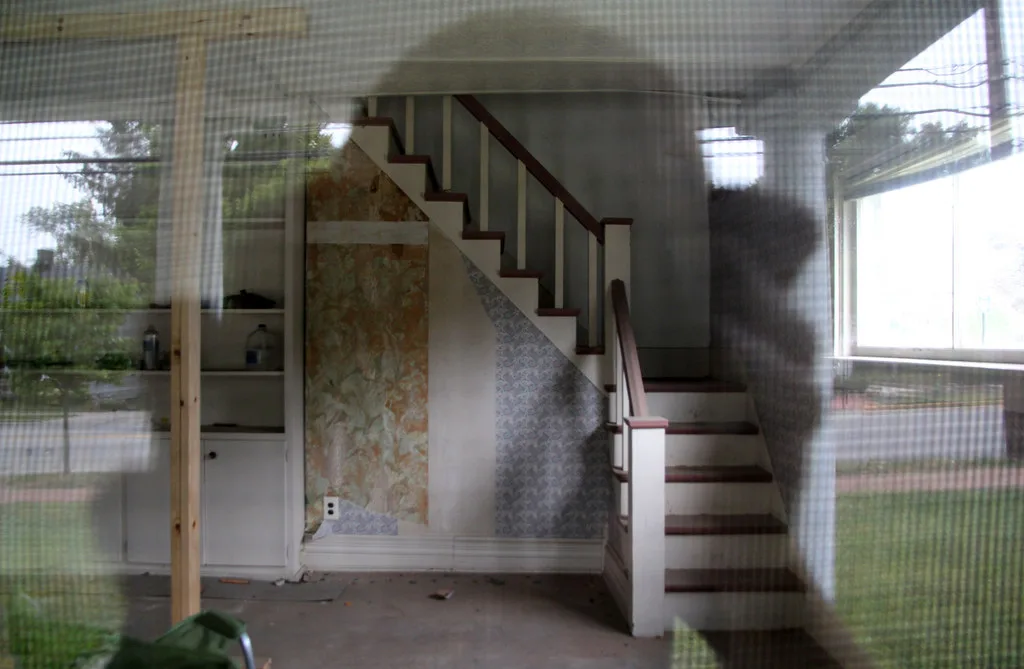 Staircase in an empty room with damaged wallpaper, seen through a window.