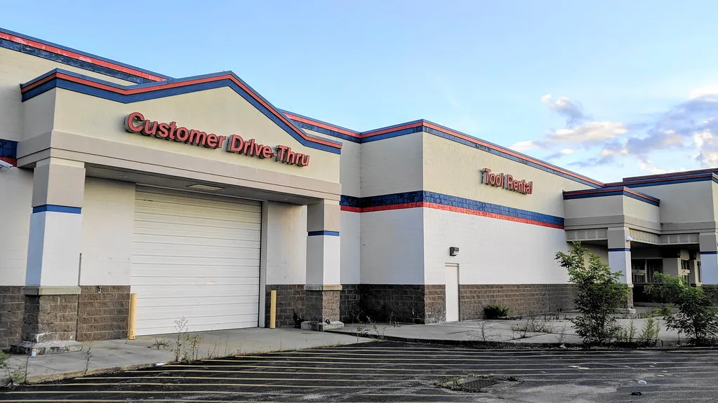 Former store with closed doors and a cracked parking area overgrown with shrubs.