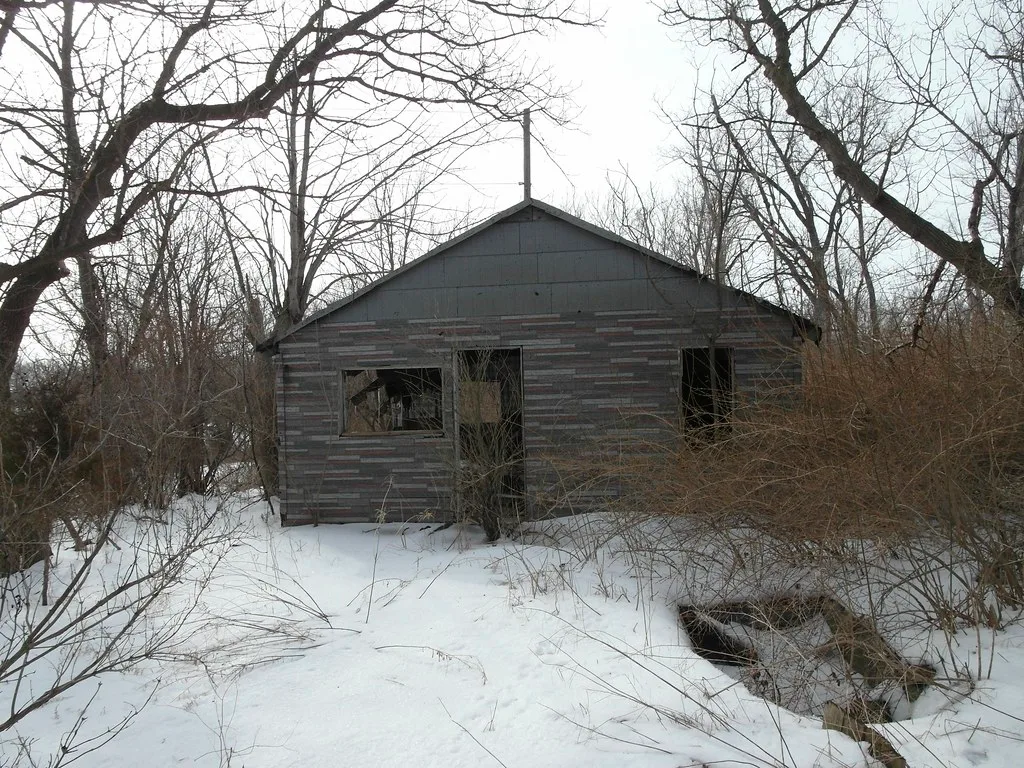 Abandoned cabin with broken windows among bare trees and snow.