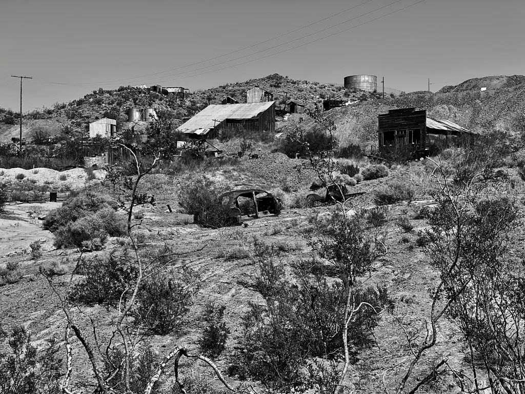 Black-and-white view of abandoned wooden buildings, tanks and vehicle shells on rocky scrub-covered ground.