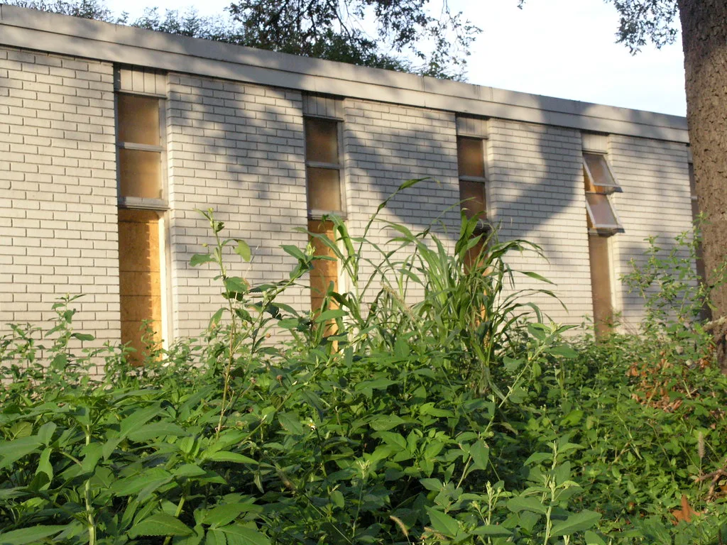 Façade abandonnée de briques claires aux fenêtres étroites condamnées et châssis penché derrière de hautes plantes.