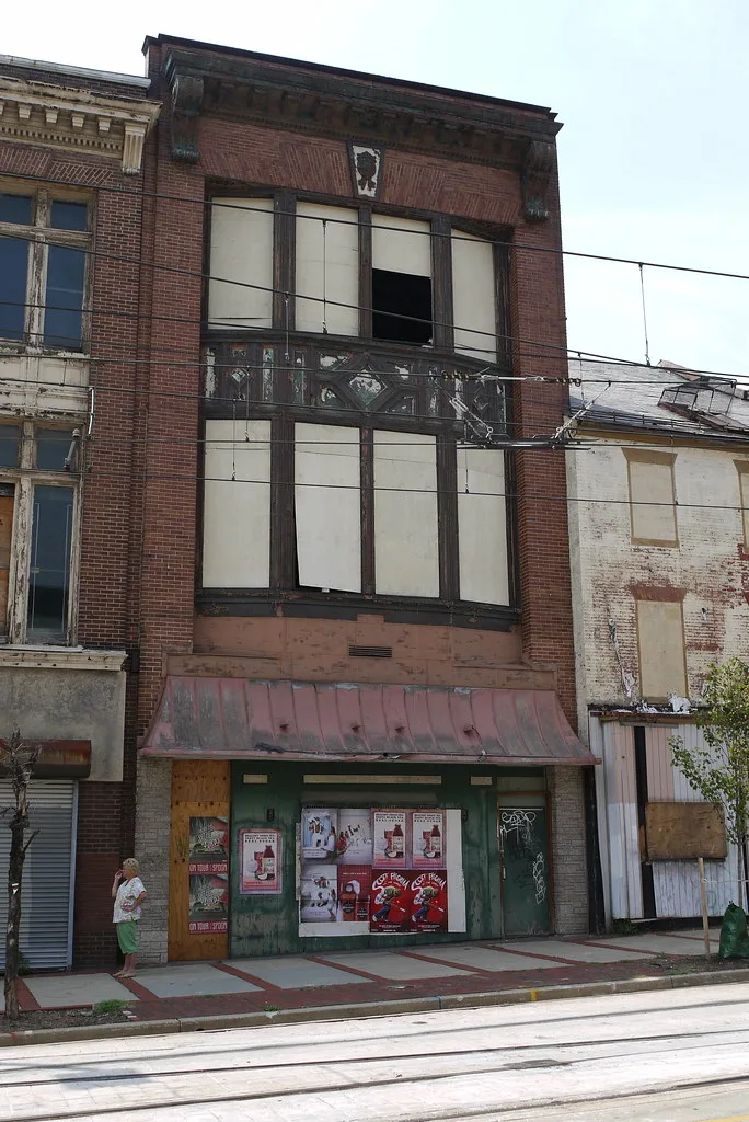 Abandoned brick facade with large boarded or empty bays, a deteriorated canopy and a former shopfront covered in posters.