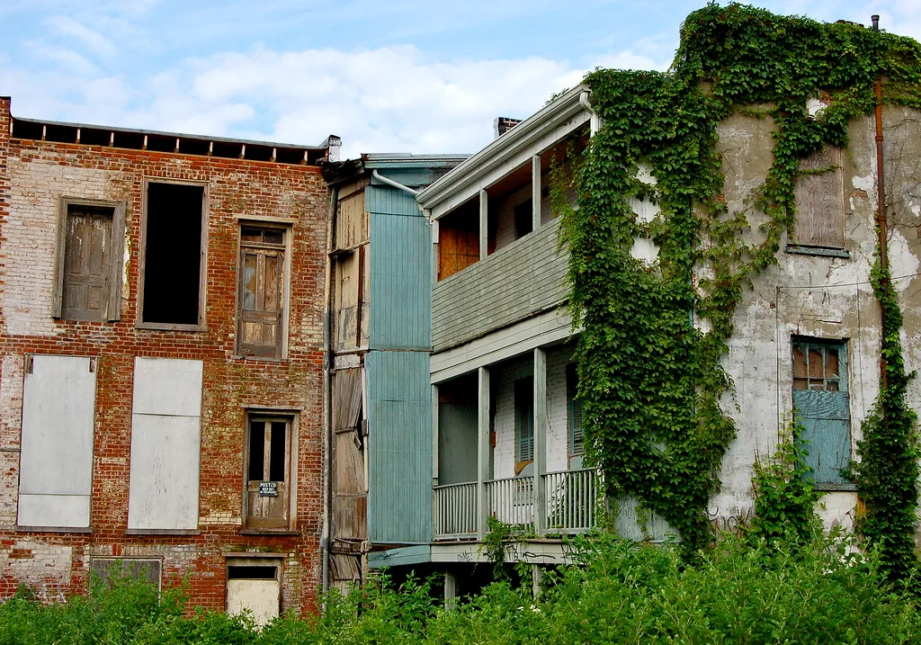 Rear of abandoned brick and wooden buildings, with empty or boarded windows and ivy-covered balconies.