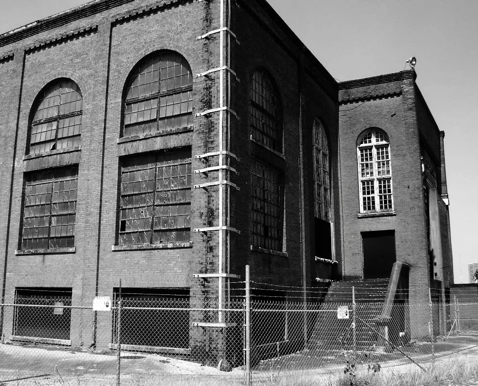 Usine de briques abandonnée en noir et blanc, aux hautes fenêtres cintrées brisées derrière un grillage.