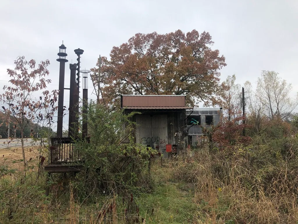 Small abandoned installation with a broken-windowed building and metal structures amid tall brush.