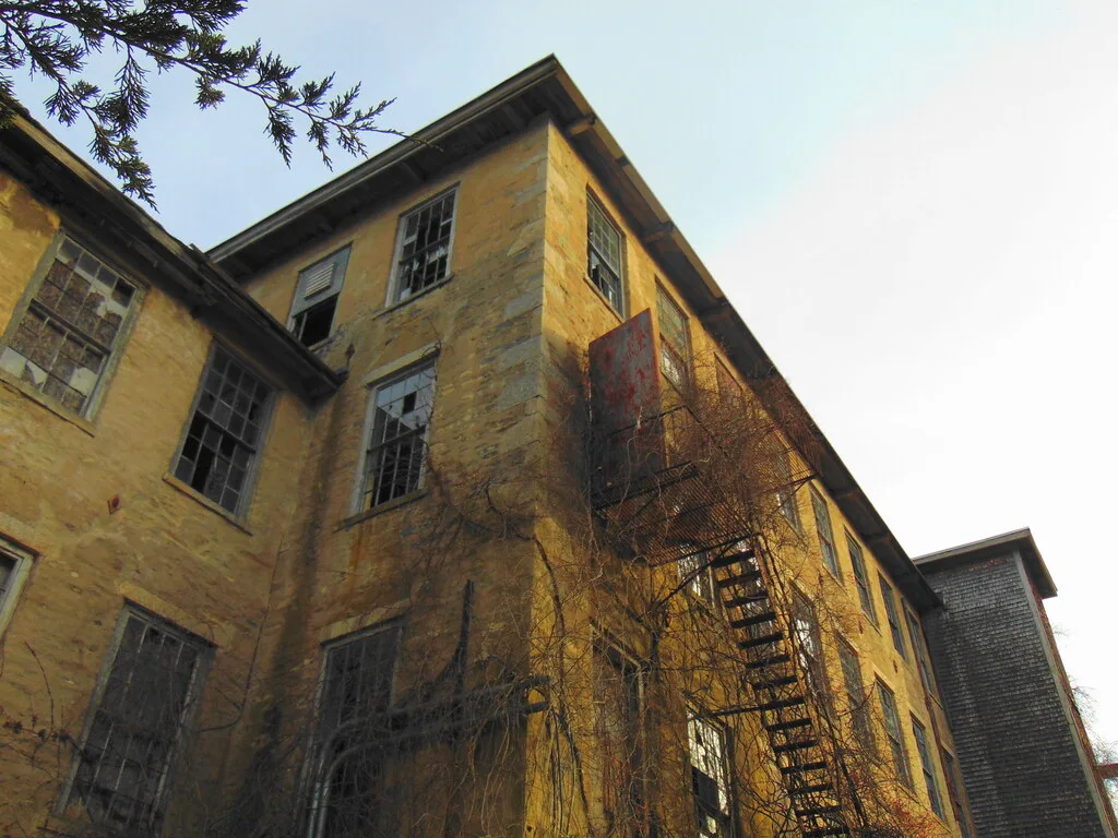Ancien bâtiment industriel jaune aux fenêtres brisées et à l’escalier métallique extérieur.