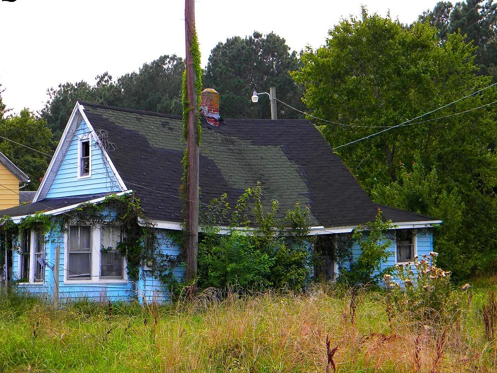 Maison bleue délaissée avec une haute toiture sombre et des arbustes envahissant le porche.