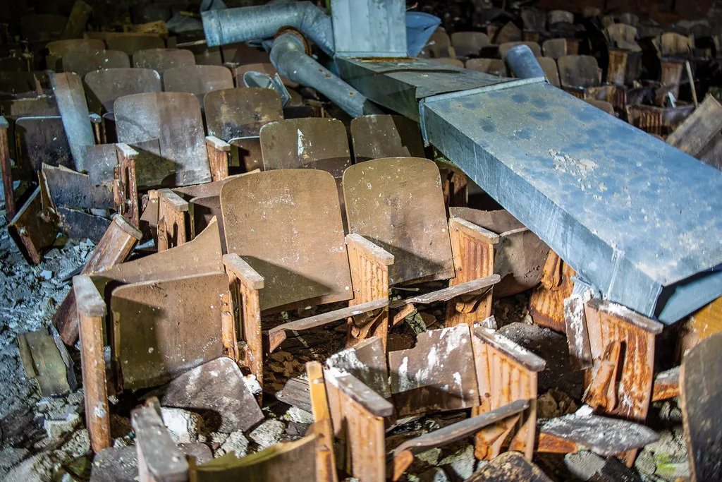 Rows of deteriorated wooden seats beneath fallen ventilation ducts.
