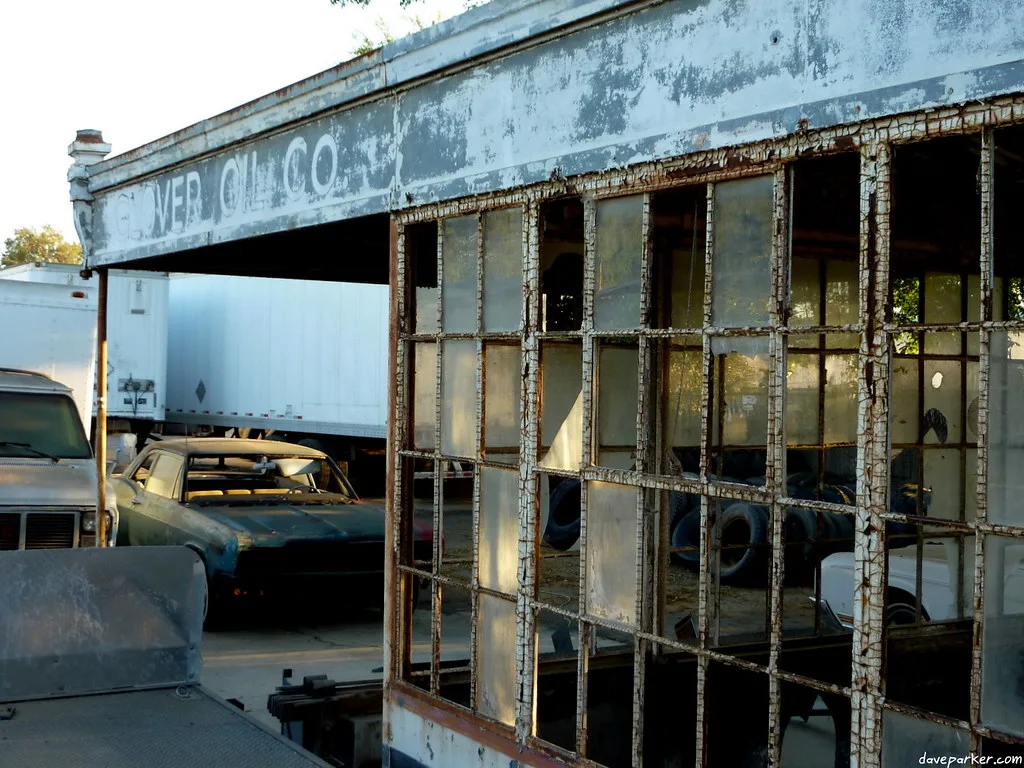 Old abandoned garage with large shattered windows and faded signage, containing tires and deteriorated cars.