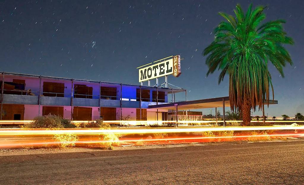 Abandoned two-storey motel with boarded windows beneath a sign, beside a palm tree and nighttime light trails.