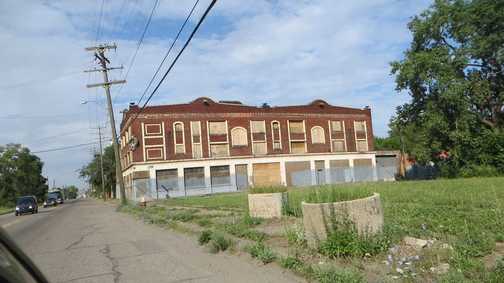 Two-storey red brick building with every opening boarded beside an overgrown plot.