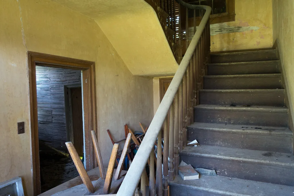 Dusty wooden staircase beside overturned chairs and walls stripped of plaster.