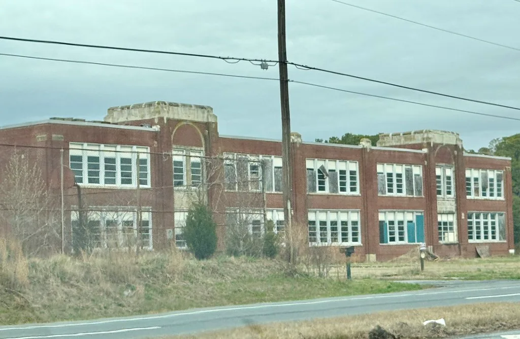 Old brick school with damaged windows behind tall grass.