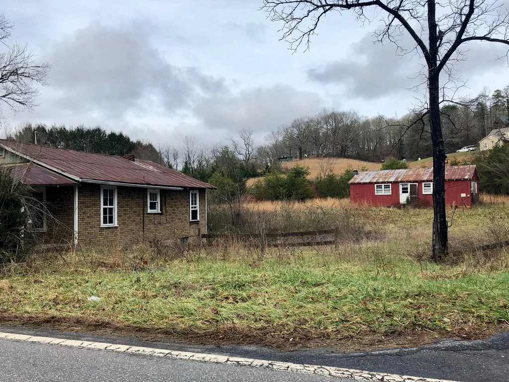 Deux petites maisons aux toits rouillés dans un terrain envahi d’herbes hautes.