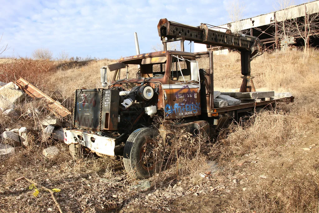 Camion-grue rouillé dans les herbes hautes devant une structure industrielle dégradée.