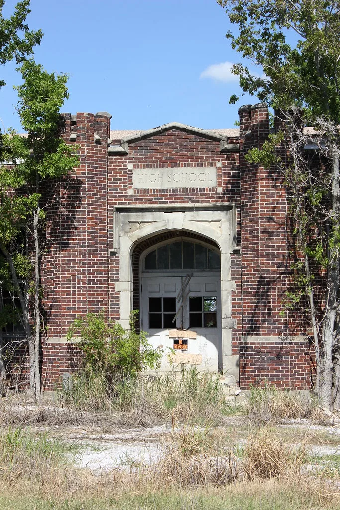 Entrée de brique d’une ancienne école, porte abîmée et partiellement obturée derrière les broussailles.