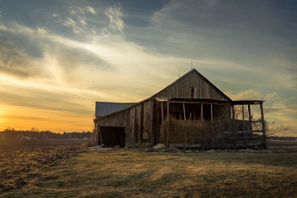Old wooden barn with a deteriorated porch beneath a sunset sky.