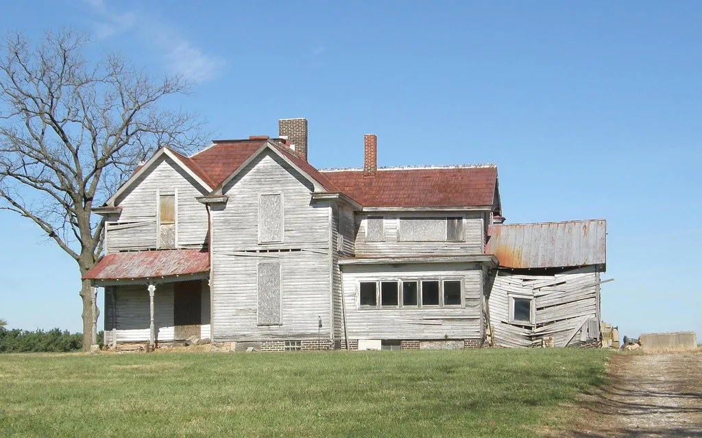 Large abandoned wooden house with boarded windows and a warped extension beneath red roofs.