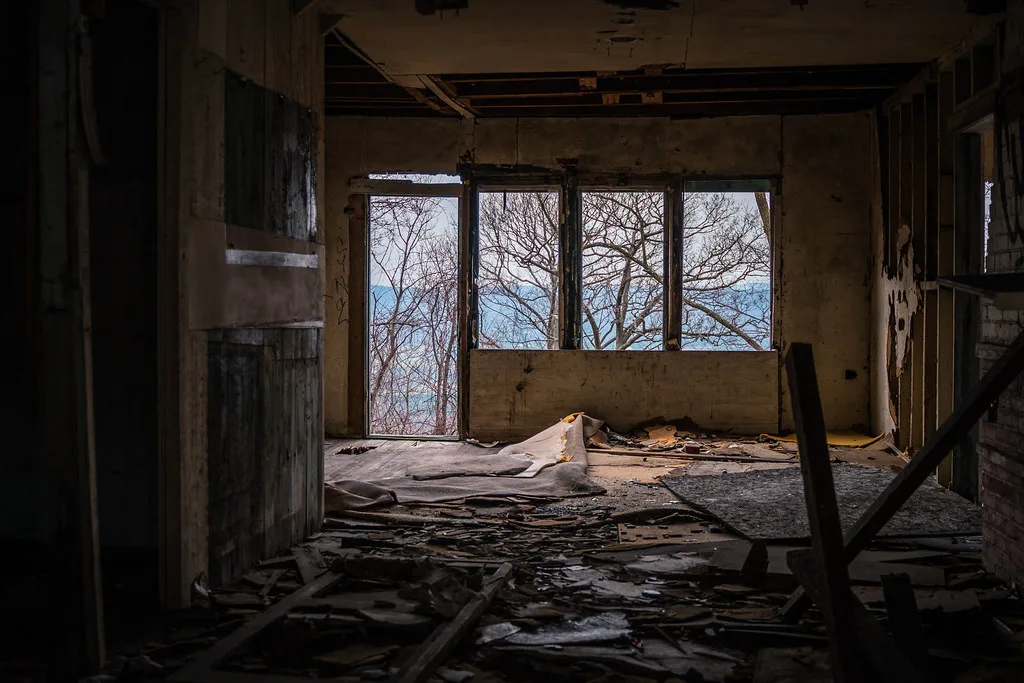 Abandoned room with exposed walls and ceiling, strewn with boards and coverings, with large openings onto trees and water.