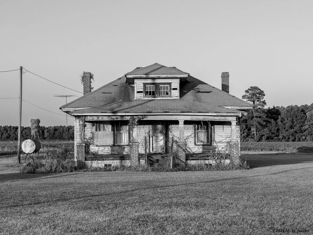 Old house with an overgrown porch and boarded windows, in black and white.