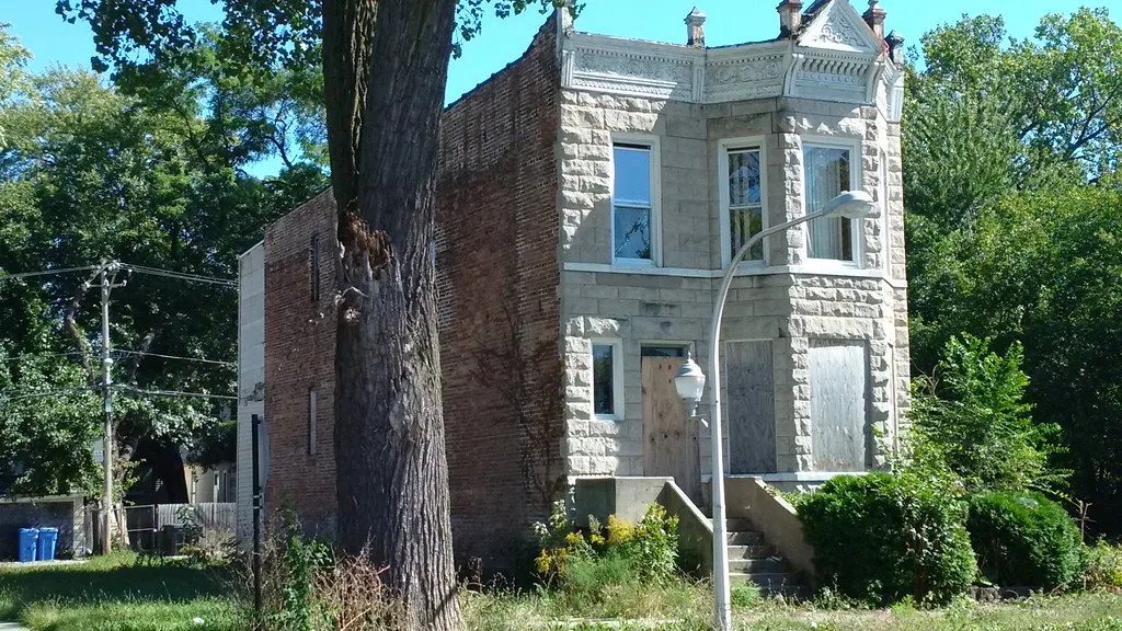 Maison étroite à façade de pierre, porte et fenêtre barricadées derrière des herbes hautes.