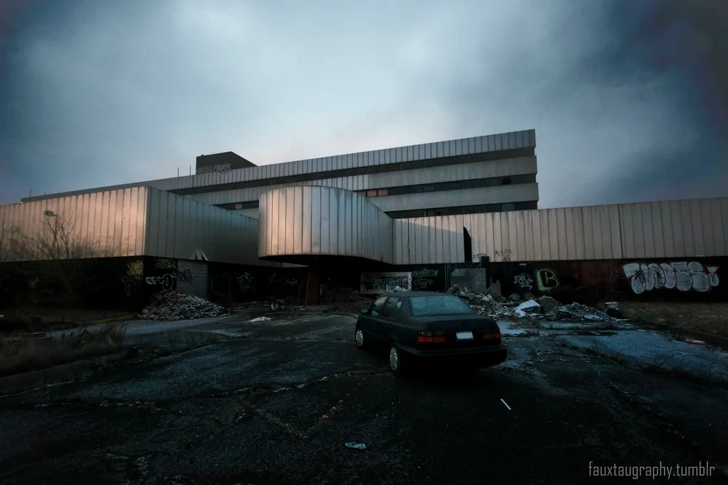Large abandoned building with metal-clad volumes before a deteriorated forecourt strewn with rubble and a car.