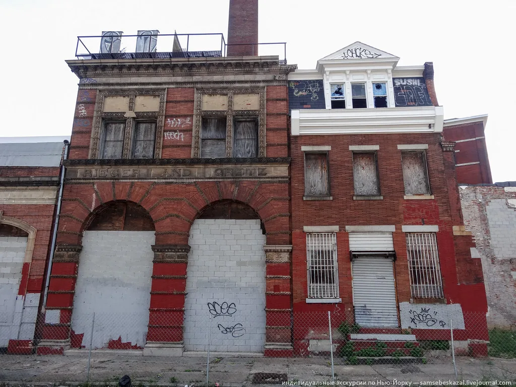 Red-brick industrial facades with walled-up arches and boarded windows beneath a chimney.