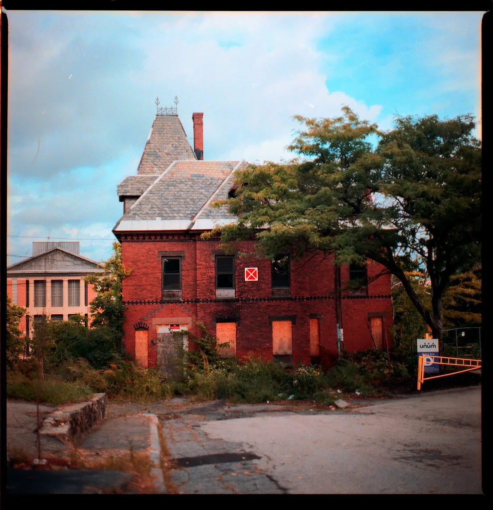 Red brick house with boarded windows behind dense vegetation.