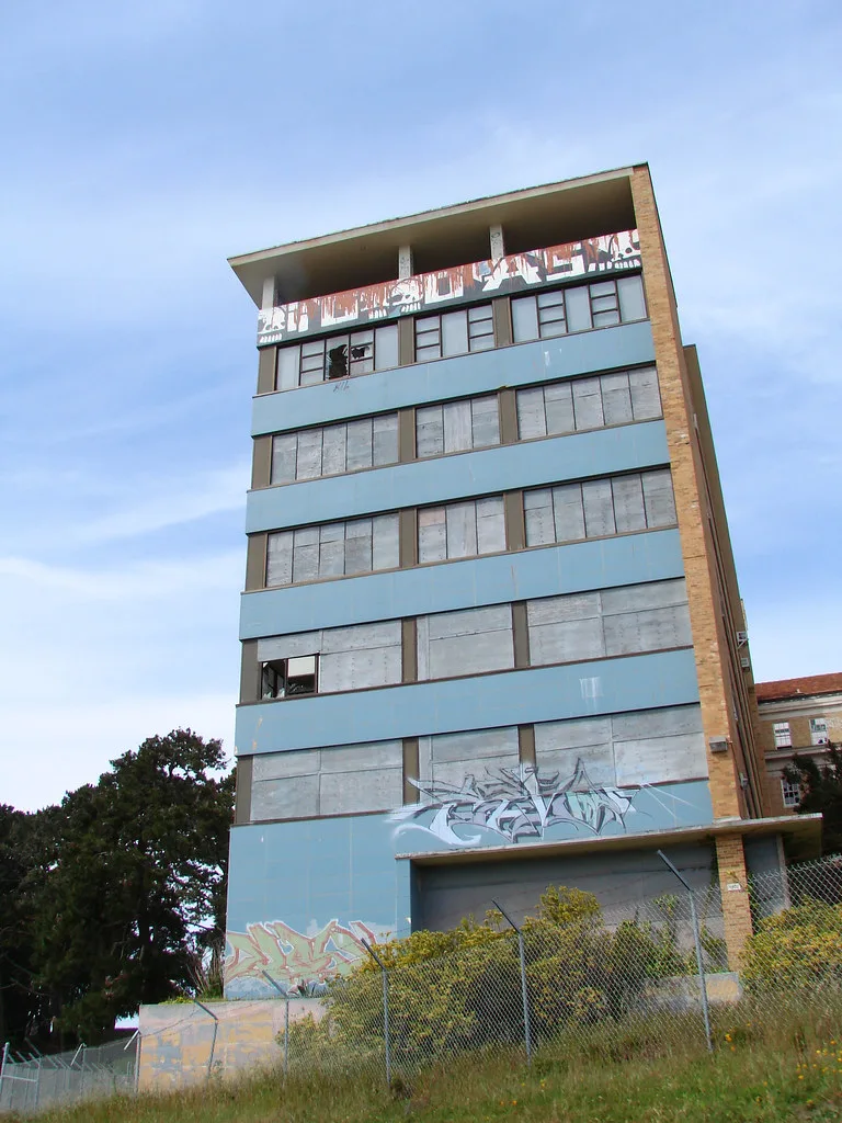 Pale blue building with rows of boarded windows and graffiti-covered walls behind a fence.