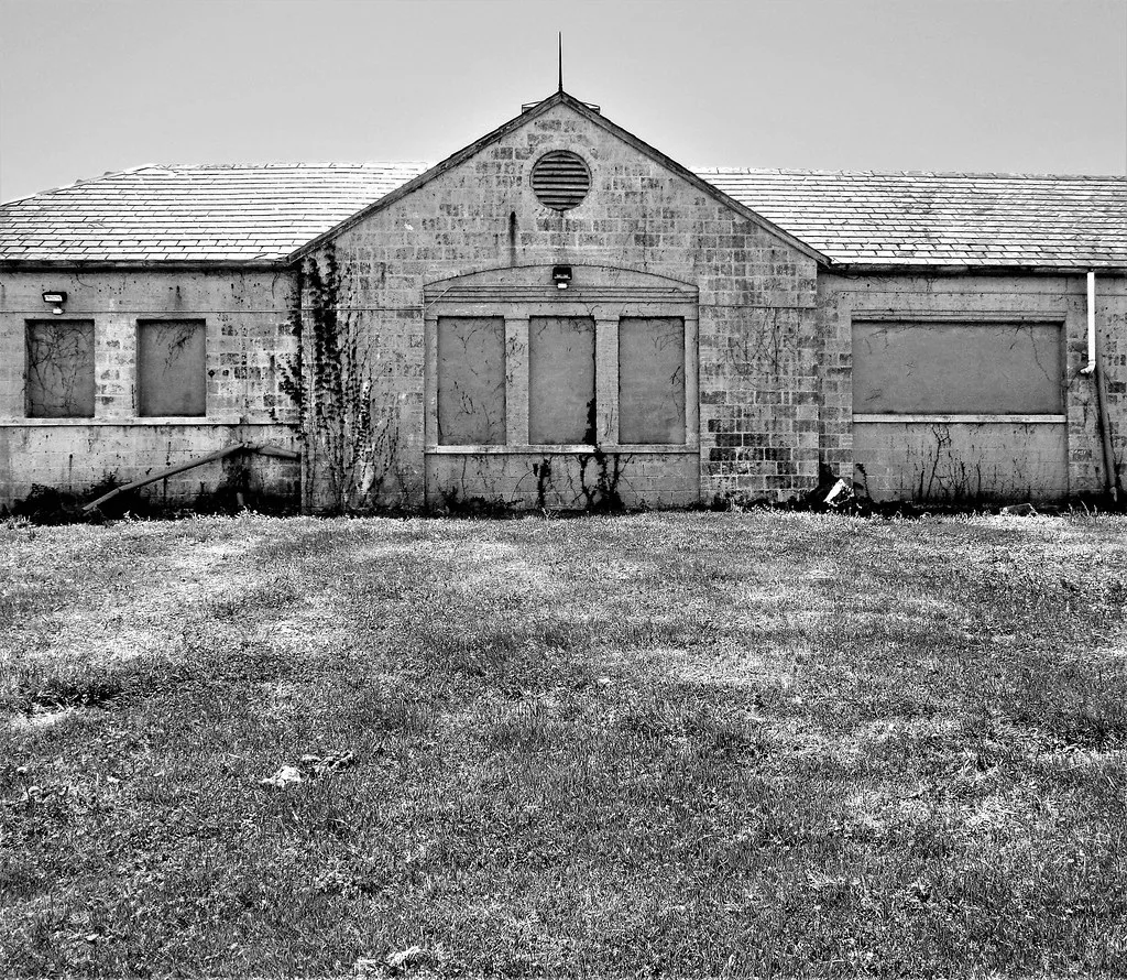 Low building with boarded openings, in black and white.