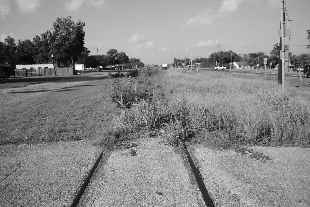 Old railway track disappearing into tall grass beyond a paved crossing.