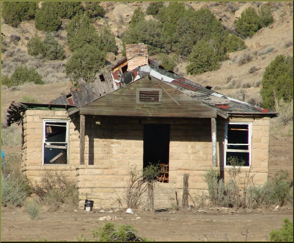 Beige stone house with a collapsed roof and open doorway on an arid slope.