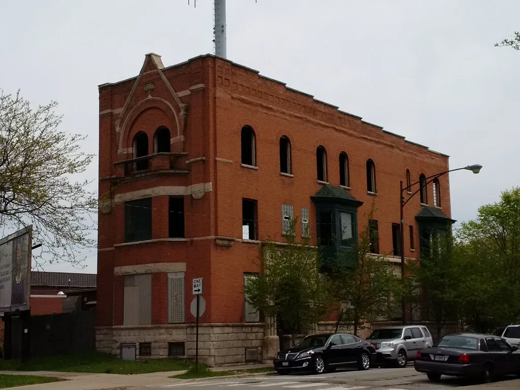 Red brick building with empty window openings and sealed entrances behind trees.
