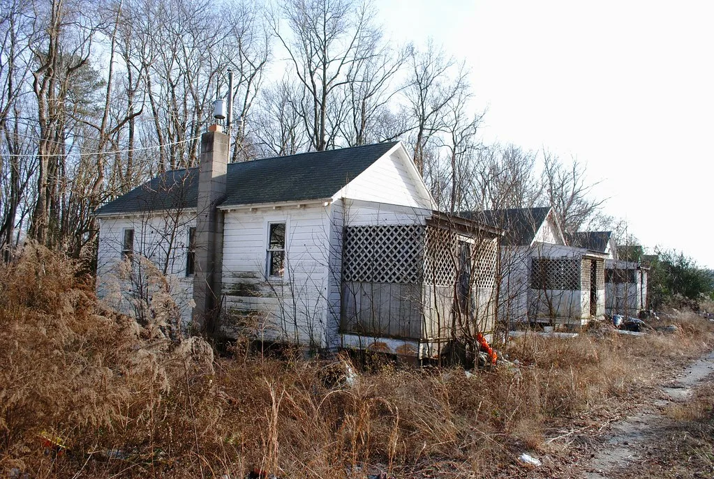 Row of neglected small white houses with broken windows and porches amid dry weeds.