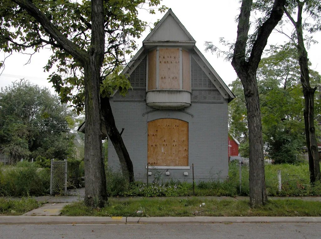 Small grey house with boarded windows between two trees.