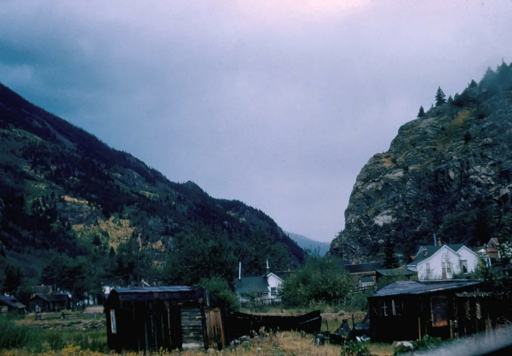 Abandoned wooden shacks with collapsed roofs and missing walls in a mountain valley.