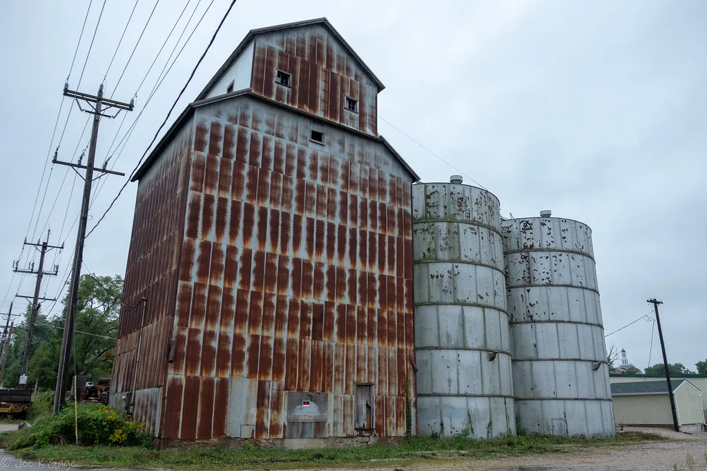 Ancien élévateur métallique rouillé accolé à deux silos blancs écaillés.
