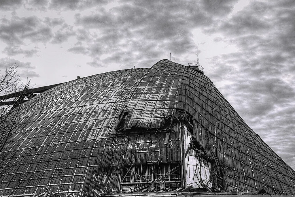 Curved roof of a large corroded hangar torn open at one corner to expose its framework.