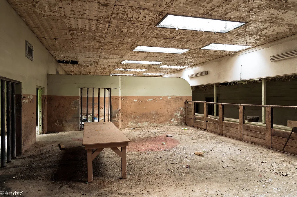Salle abandonnée éclairée par des lanterneaux, avec table de bois, cloisons éventrées et plafond fortement écaillé.